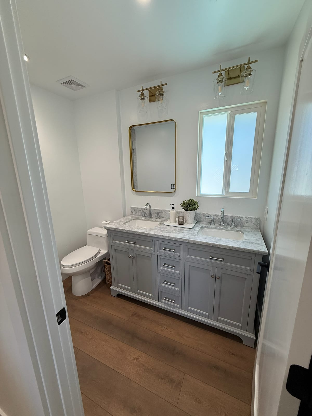 A gray double vanity with a stone top and a framed mirror, set on new wood flooring.