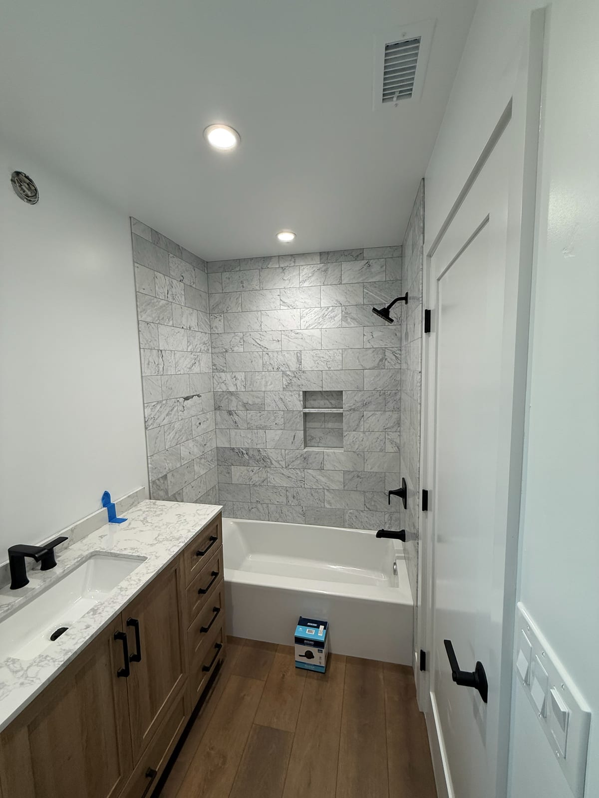 A full bath in marble subway tile: a tub and shower behind glass, matte black fixtures, and a wood vanity on new plank flooring.