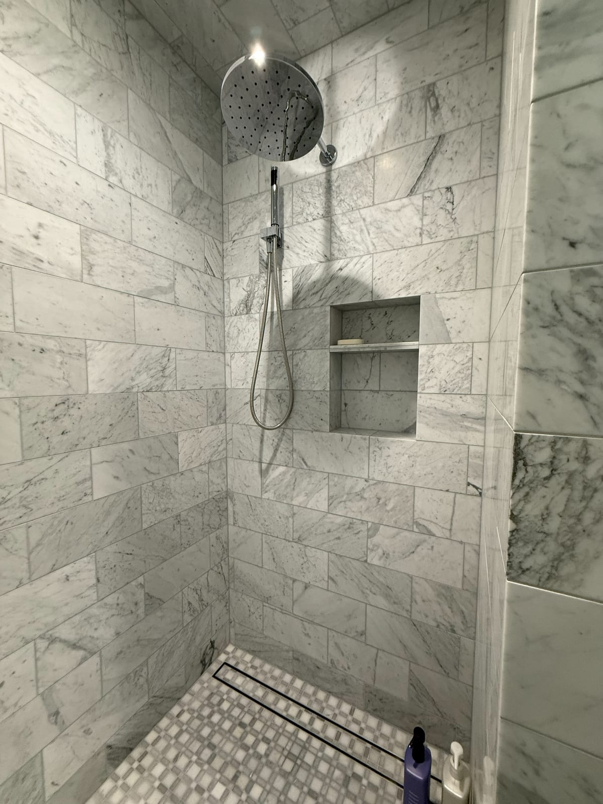 A walk-in shower wrapped in Carrara marble with a rainfall head, a built-in niche, and a pebble floor. Tile this size only lines up when the layout is set before the first cut.